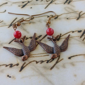 May include: A pair of earrings featuring a copper-colored bird design with a red bead accent.
