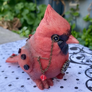 May include: A red cardinal figurine with a steampunk design. The cardinal has a monocle, a chain, buttons, and a gear. The figurine is sitting on a white and black polka dot surface.
