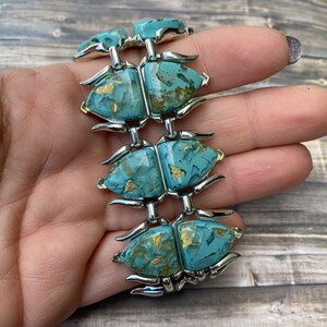 May include: A vintage bracelet with six turquoise beetle-shaped links, each with gold flecks, joined by silver-toned metal. The bracelet is held in a hand, highlighting its intricate design and colour palette.
