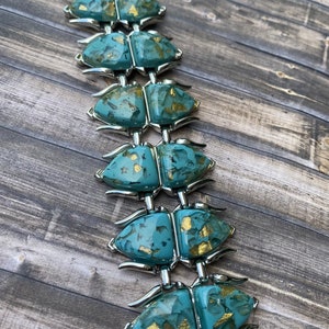 May include: A vintage bracelet with turquoise beetle-shaped links, adorned with gold flecks, linked by silver-toned metal. The bracelet is displayed on a wooden surface, highlighting its unique design and colour palette.