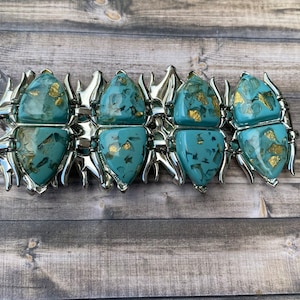 May include: A silver bracelet with turquoise beetle-shaped links, each with gold flecks. The bracelet is hinged and displayed on a wooden surface. The links are connected by silver metal. The bracelet is a statement piece.
