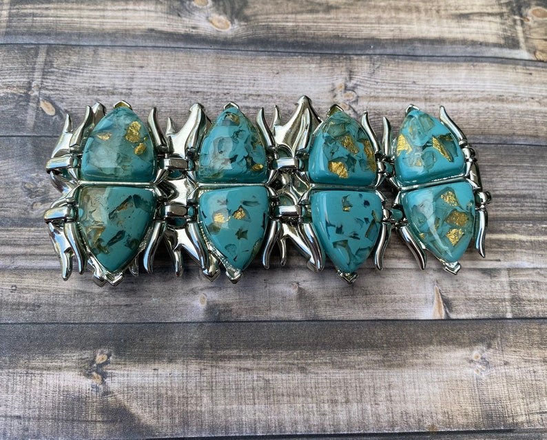 May include: A silver bracelet with turquoise beetle-shaped links, each with gold flecks. The bracelet is hinged and displayed on a wooden surface. The links are connected by silver metal. The bracelet is a statement piece.