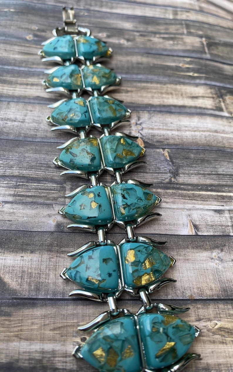 May include: A vintage bracelet with turquoise-coloured, abstract links with gold flecks, joined by silver-toned metal. The bracelet is displayed on a wooden surface, highlighting its unique design and colour palette.