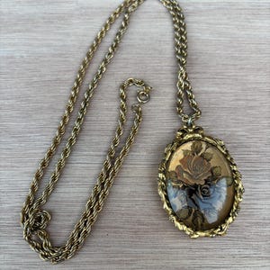 May include: A gold-tone rope chain necklace featuring an oval pendant with a textured, ornate frame. The pendant contains a central glass or resin insert displaying a detailed illustration of a blooming rose with leaves in muted brown and tan tones.