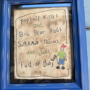 Pode incluir: Uma obra de arte emoldurada com uma moldura de madeira azul. A obra apresenta texto manuscrito: "Popsicle Kisses and Big Bear Hugs, Superman Trains and Jars Full of Bugs." Uma pequena figura de desenho animado é desenhada no canto inferior direito.