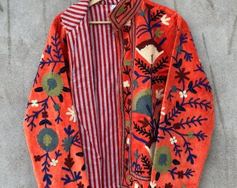 Könnte beinhalten: Eine leuchtend orangefarbene Jacke mit aufwendigen Blumenstickereien in Blau-, Grün- und Weißtönen. Das Kleidungsstück hat einen Umlegekragen und eine offene Vorderseite, die ein rot-weiß gestreiftes Innenfutter zum Vorschein bringt.