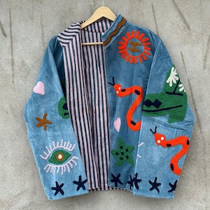 May include: A light blue jacket featuring colorful embroidered patches including a sun, snakes, an eye, stars, and hearts. The interior lining is visible and displays a vertical red, white, and black striped pattern. The jacket hangs on a wooden hanger.