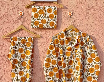 Könnte beinhalten: Ein weißes Pyjama-Set mit einem sich wiederholenden Muster aus orangefarbenen Ringelblumen mit grünen Stielen und Blättern. Das Set besteht aus einem langärmeligen Hemd mit Knopfleiste, Kragen und Brusttasche sowie einer passenden Hose. Ein Schild zeigt ETSY SHOP SEHANDICRAFTS.