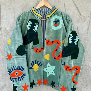 May include: A light green velvet jacket with colorful embroidered designs. The jacket features a sun, snakes, boots, stars, and an eye. The collar has yellow and blue stripes. The jacket is hanging on a wooden hanger.