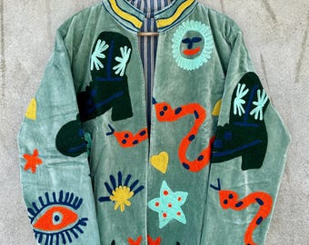 Könnte beinhalten: Hellgrüne Samtjacke mit bunten Stickereien. Die Jacke hat eine Sonne, Schlangen, Stiefel, Sterne und ein Auge. Der Kragen hat gelbe und blaue Streifen. Die Jacke hängt an einem Holzkleiderbügel.