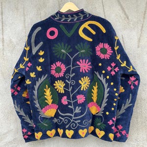 May include: A dark blue jacket featuring vibrant, multi-colored embroidery. The back displays the word LOVE in large letters above a floral pattern with pink, yellow, and green flowers, leaves, and small yellow hearts along the bottom hem.
