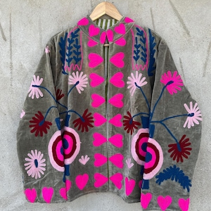 以下が含まれることがあります： 鮮やかな花の刺繍が施されたグレーのベルベットジャケット。ピンクのハート、花、青い葉のようなデザインが特徴です。裏地は緑と白のストライプです。木製のハンガーにかかっています。
