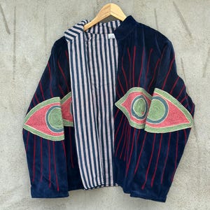 May include: A dark blue velvet jacket featuring large embroidered eye motifs on the sleeves and front panels. The eyes are rendered in shades of green, pink, and blue with red radiating lines. The interior is lined with a white and blue striped fabric.