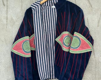 May include: A dark blue velvet jacket featuring large embroidered eye motifs on the sleeves and front panels. The eyes are rendered in shades of green, pink, and blue with red radiating lines. The interior is lined with a white and blue striped fabric.