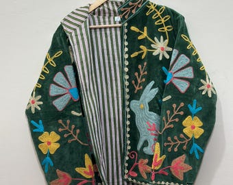 Könnte beinhalten: Eine dunkelgrüne Jacke mit bunter Stickerei, die einen Hasen und verschiedene Blumenmuster zeigt. Die Jacke hat einen Stehkragen und ist mit einem weiß-grün gestreiften Stoff gefüttert. Sie hängt an einem Holzkleiderbügel vor einer schlichten Wand.