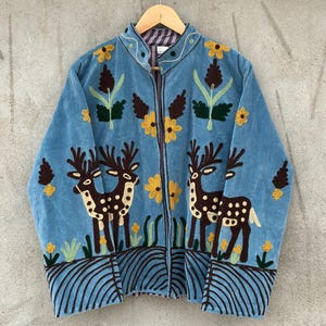 May include: A light blue zip-up jacket featuring dark brown deer silhouettes with white spots, yellow flower embroidery, and dark brown curved line patterns along the hem. The jacket has a stand-up collar and long sleeves with a striped inner lining.