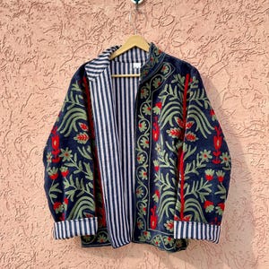 May include: A dark blue jacket featuring intricate floral embroidery in shades of green and red. The garment has a stand-up collar, long sleeves with folded cuffs, and an interior lining with a vertical white and dark blue striped pattern.