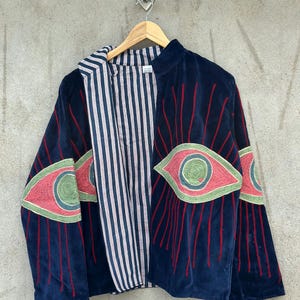 May include: A dark blue velvet jacket featuring large embroidered eye motifs in pink, green, and blue on the sleeves and front panels. The jacket has a high collar and is lined with a white fabric featuring thin vertical navy blue stripes.