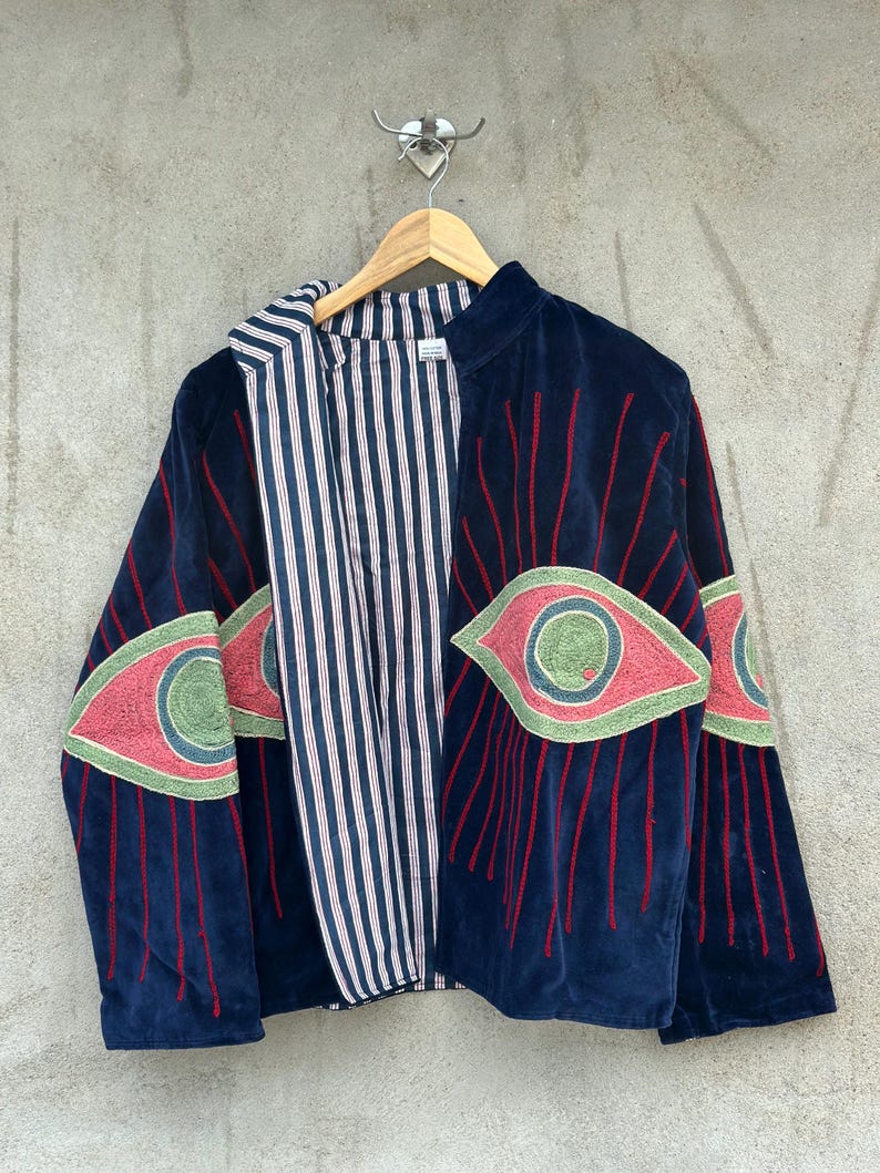 May include: A dark blue velvet jacket featuring large embroidered eye motifs in pink, green, and blue on the sleeves and front panels. The jacket has a high collar and is lined with a white fabric featuring thin vertical navy blue stripes.