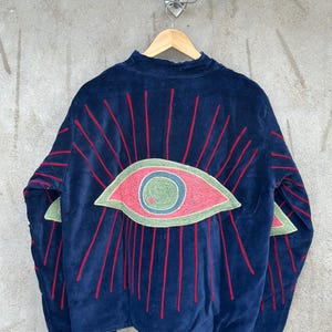 May include: A dark blue velvet jacket featuring a large embroidered eye design on the back. The eye is rendered in shades of pink, green, and blue, surrounded by radiating red embroidered lines that extend across the back and onto the sleeves.