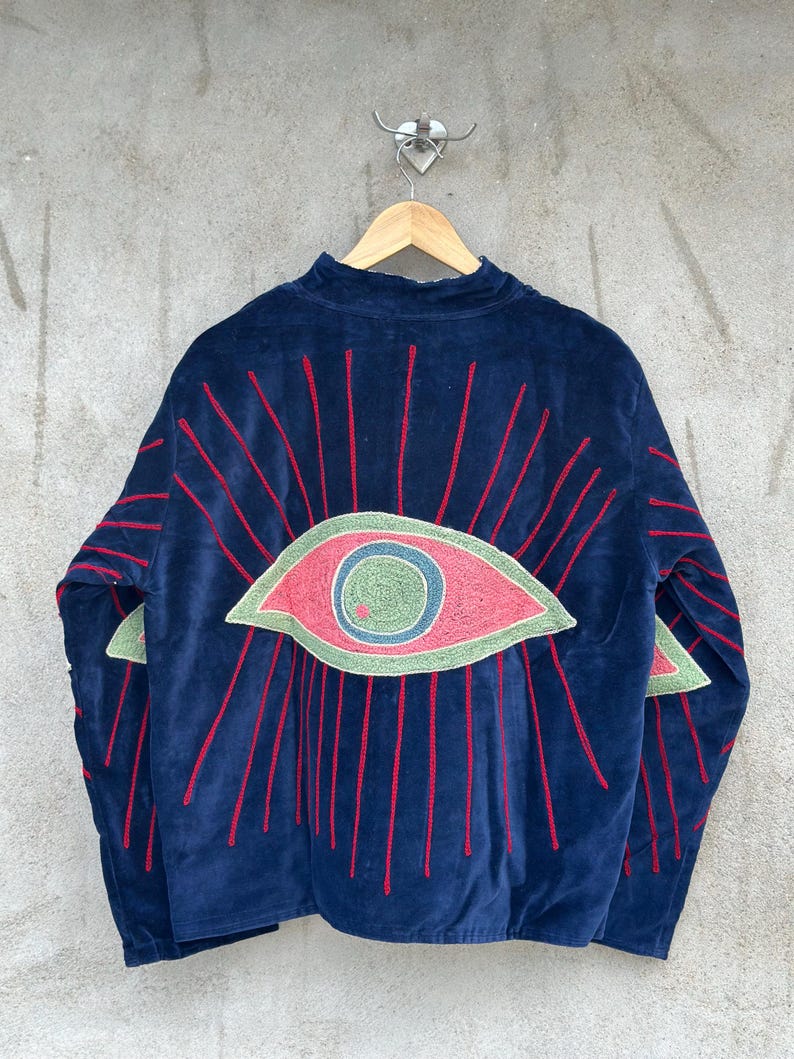 May include: A dark blue velvet jacket featuring a large embroidered eye design on the back. The eye is rendered in shades of pink, green, and blue, surrounded by radiating red embroidered lines that extend across the back and onto the sleeves.