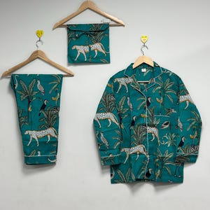 May include: Teal pyjama set with a jungle print featuring leopards, toucans, and palm trees. The set includes a button-down shirt, trousers, and a matching travel pouch, all trimmed with white piping. The pyjamas are hanging on wooden hangers.