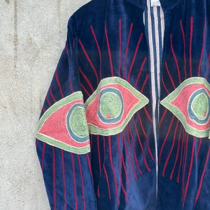 May include: A dark blue velvet jacket featuring large embroidered eye motifs in shades of pink, green, and blue. The design includes radiating red stitched lines extending from the eyes. The interior lining is visible, showing a white and red striped pattern.