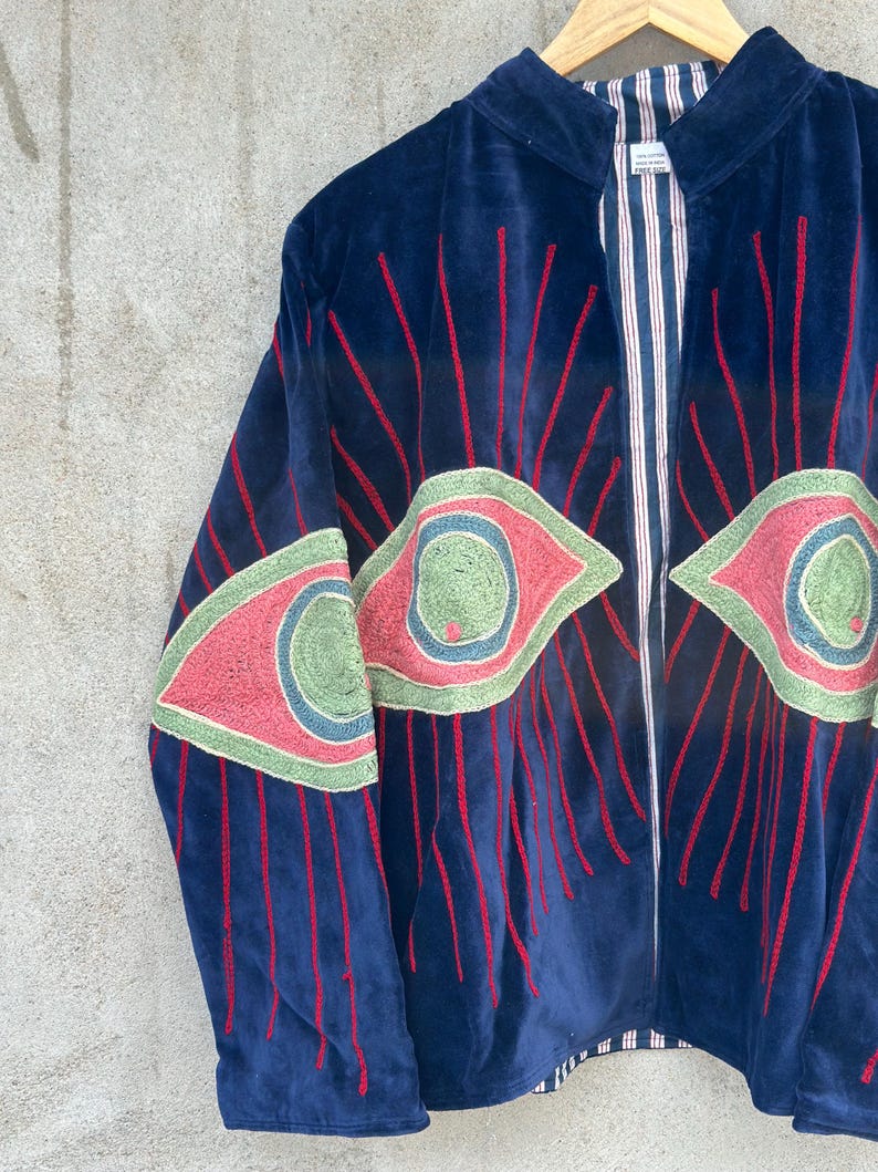 May include: A dark blue velvet jacket featuring large embroidered eye motifs in shades of pink, green, and blue. The design includes radiating red stitched lines extending from the eyes. The interior lining is visible, showing a white and red striped pattern.