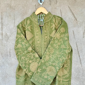 Op de afbeelding: Een groene jas met ingewikkelde bloemenborduursels in een lichtere groentint. De jas heeft een hoge kraag en lange mouwen. Hij hangt aan een houten kleerhanger tegen een neutraal gekleurde muur.