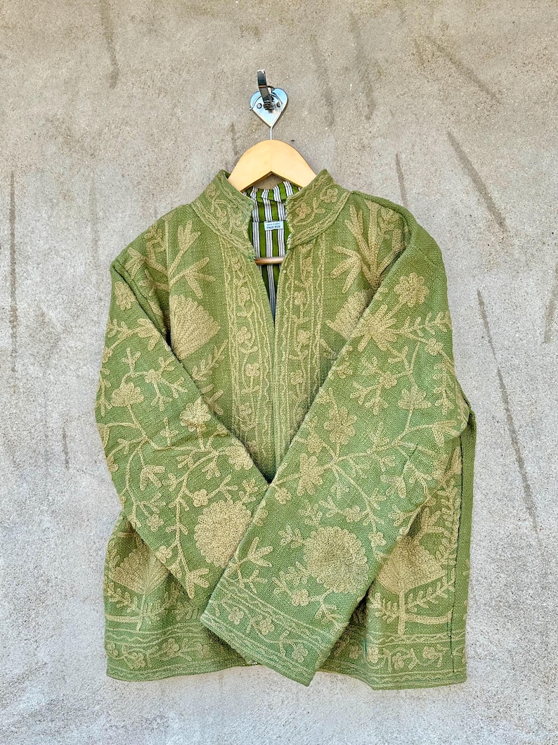 Op de afbeelding: Een groene jas met ingewikkelde bloemenborduursels in een lichtere groentint. De jas heeft een hoge kraag en lange mouwen. Hij hangt aan een houten kleerhanger tegen een neutraal gekleurde muur.