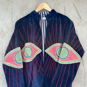 May include: A navy blue velvet jacket featuring large embroidered eye motifs on the front panels. The eyes are detailed with pink, light green, and blue thread, surrounded by radiating red embroidered lines. The interior lining has a white and blue stripe.