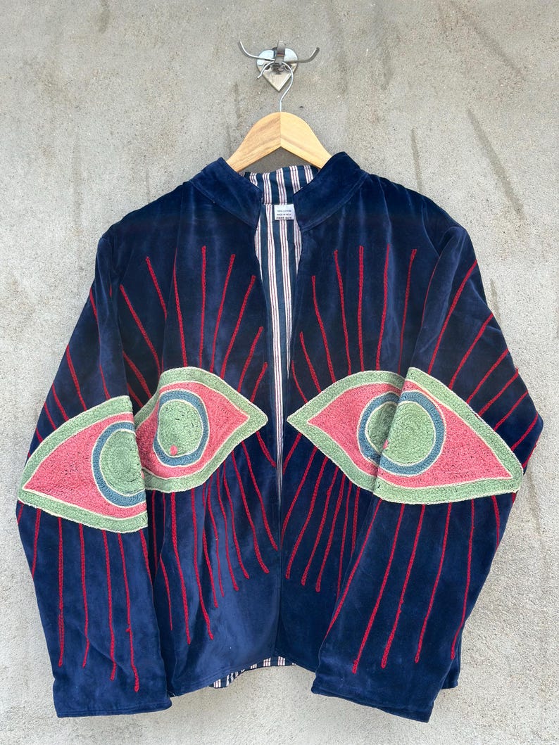 May include: A navy blue velvet jacket featuring large embroidered eye motifs on the front panels. The eyes are detailed with pink, light green, and blue thread, surrounded by radiating red embroidered lines. The interior lining has a white and blue stripe.