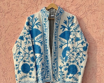 Könnte beinhalten: Eine langärmelige Jacke aus cremefarbenem Stoff mit leuchtend blauer Blumenstickerei auf der Vorderseite und den Ärmeln. Das Kleidungsstück hat einen Stehkragen, eine offene Vorderseite und ein sichtbares dunkelgrau-weiß gestreiftes Innenfutter.