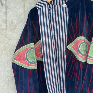 May include: A dark blue velvet jacket featuring large embroidered eye motifs on the sleeves and front. The embroidery uses shades of pink, green, and blue with red radiating lines. The jacket has a white and navy blue vertical striped lining.