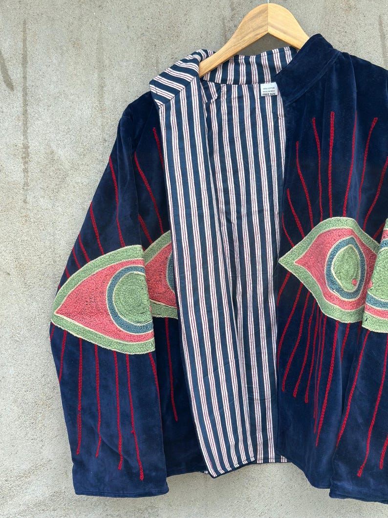 May include: A dark blue velvet jacket featuring large embroidered eye motifs on the sleeves and front. The embroidery uses shades of pink, green, and blue with red radiating lines. The jacket has a white and navy blue vertical striped lining.