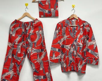 May include: A red pajama set featuring a repeating pattern of abstract figures in white and black. The set includes trousers, a long-sleeved button-down shirt, and a matching scarf. The pajamas are displayed on wooden hangers against a white backdrop.