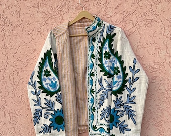 Könnte beinhalten: Eine langärmelige Jacke mit cremefarbener Basis und auffälligen grünen und blauen Blumen- und Paisley-Stickereien. Das Innenfutter zeigt ein hellbraunes und weißes vertikales Streifenmuster. Die Jacke verfügt über einen hohen Stehkragen.