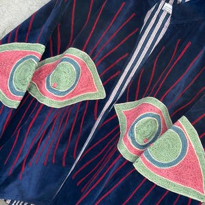 May include: A navy blue velvet jacket featuring red embroidered vertical lines and large, stylized eye-shaped patches in shades of green, pink, and blue. The jacket has a stand-up collar and a white and navy blue striped lining visible at the opening.