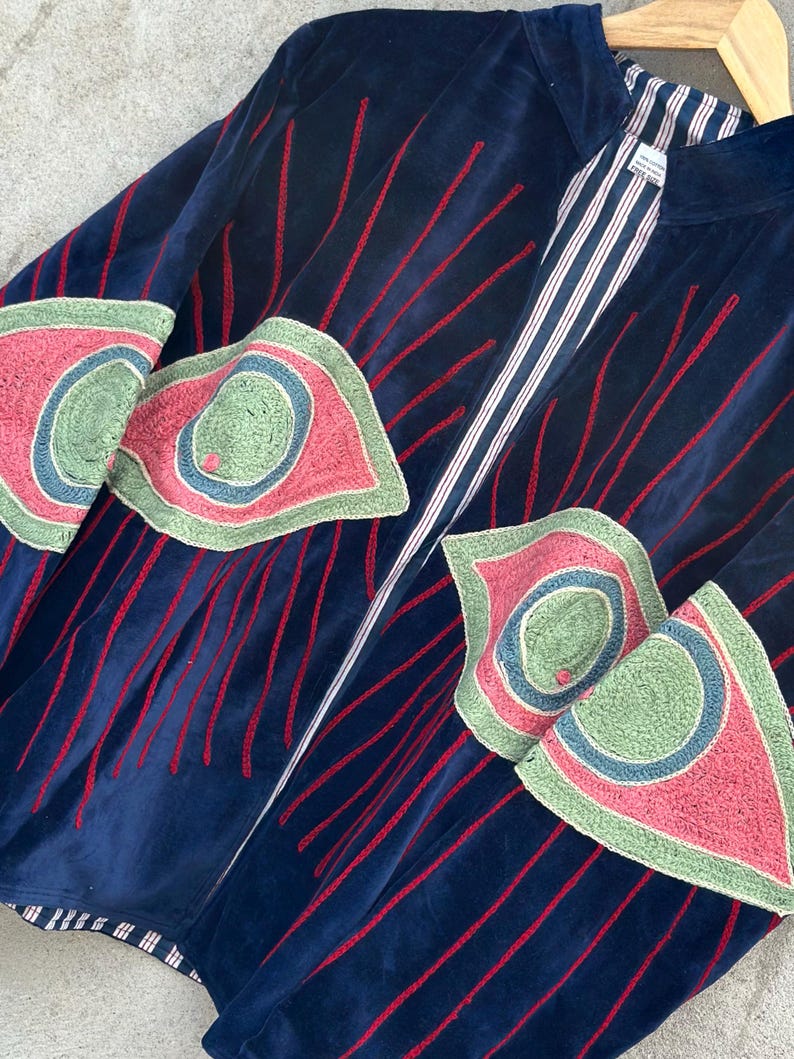 May include: A navy blue velvet jacket featuring red embroidered vertical lines and large, stylized eye-shaped patches in shades of green, pink, and blue. The jacket has a stand-up collar and a white and navy blue striped lining visible at the opening.