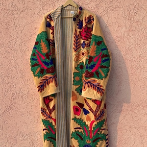 Puede incluir: Un abrigo largo abierto por delante en color beige claro con bordados florales y de hojas en tonos vibrantes de verde, rojo, azul y naranja. El forro interior muestra un sutil patrón de rayas verticales en gris y blanco.