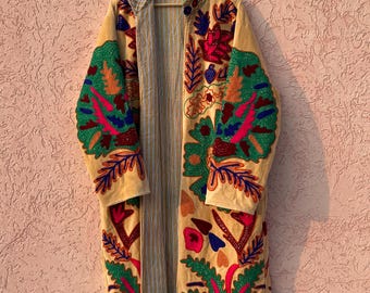 Könnte beinhalten: Ein langer, vorne offener Mantel in hellem Beige mit leuchtend bunter Blumen- und Blattstickerei in Grün, Rot, Blau und Orange. Das Innenfutter zeigt ein dezentes Muster aus vertikalen grauen und weißen Streifen.
