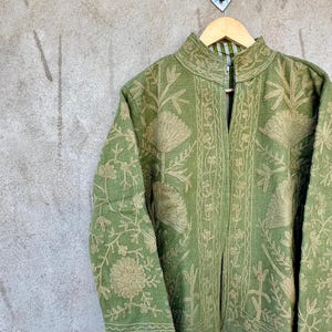 Op de afbeelding: Een groene jas met ingewikkelde bloemenborduursels in een lichtere groentint. De jas heeft een hoge kraag en lange mouwen. Hij hangt aan een houten hanger tegen een grijze, getextureerde muur.