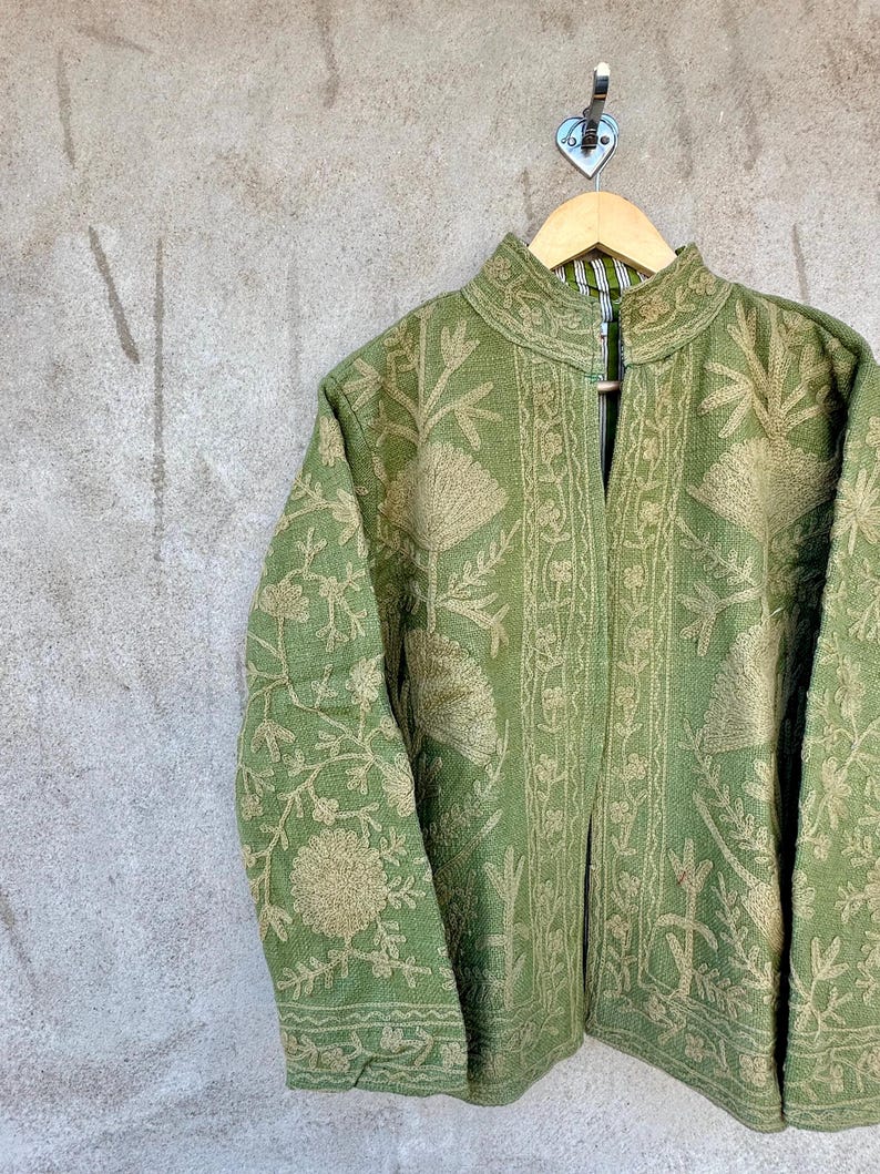 Op de afbeelding: Een groene jas met ingewikkelde bloemenborduursels in een lichtere groentint. De jas heeft een hoge kraag en lange mouwen. Hij hangt aan een houten hanger tegen een grijze, getextureerde muur.