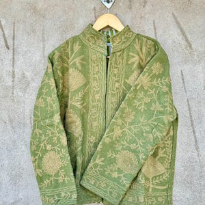 Op de afbeelding: Een groene jas met ingewikkelde bloemenborduursels in een lichtere tint groen. De jas heeft een opstaande kraag en lange mouwen. Hij hangt aan een houten hanger tegen een neutraal gekleurde muur.