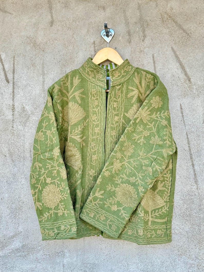 Op de afbeelding: Een groene jas met ingewikkelde bloemenborduursels in een lichtere tint groen. De jas heeft een opstaande kraag en lange mouwen. Hij hangt aan een houten hanger tegen een neutraal gekleurde muur.