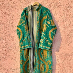 Può includere: Un cappotto a maniche lunghe con un esterno in velluto verde brillante decorato con intricati ricami color oro in motivi floreali e a foglia. La fodera interna presenta un design a righe verticali nei toni del bianco, del verde e del marrone.
