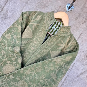 Op de afbeelding: Saliegroene jas met ingewikkelde bloemenborduurwerk. De jas heeft een opstaande kraag en een gestreepte voering. Het wordt gepresenteerd op een houten hanger met een metalen haak.