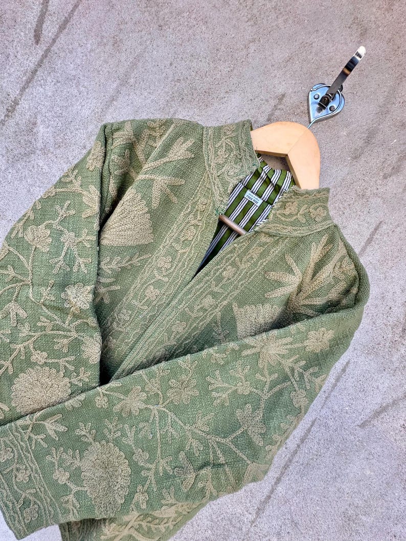 Op de afbeelding: Saliegroene jas met ingewikkelde bloemenborduurwerk. De jas heeft een opstaande kraag en een gestreepte voering. Het wordt gepresenteerd op een houten hanger met een metalen haak.
