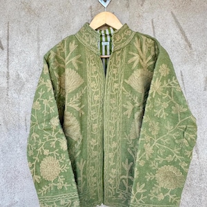 Op de afbeelding: Een groene jas met ingewikkelde bloemenborduursels in een lichtere groentint. De jas heeft een opstaande kraag en lange mouwen. Hij hangt aan een houten kleerhanger, met een gestreepte voering zichtbaar bij de kraag.