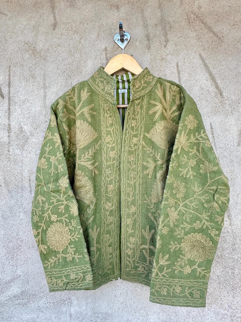Op de afbeelding: Een groene jas met ingewikkelde bloemenborduursels in een lichtere groentint. De jas heeft een opstaande kraag en lange mouwen. Hij hangt aan een houten kleerhanger, met een gestreepte voering zichtbaar bij de kraag.